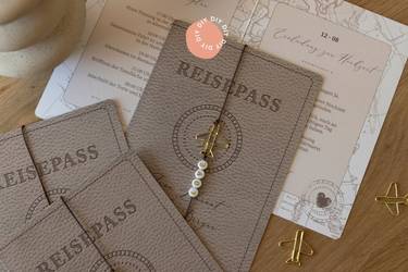  Hochzeitseinladung im Reisepass-Stil verziert mit Perlen und einer Heftklammer in Flugzeugform.