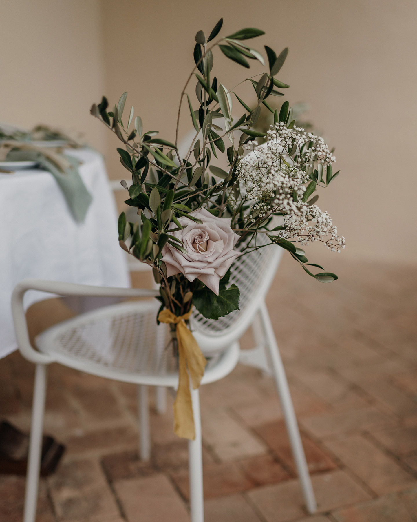 Der weiße Stuhl in Gitter-Optik steht vor dem Hochzeitstisch. Geschmückt ist er mit einem Blumenstrauß aus Olivenzweigen, Schleierkraut und einer Rose in hellem Altrosa.