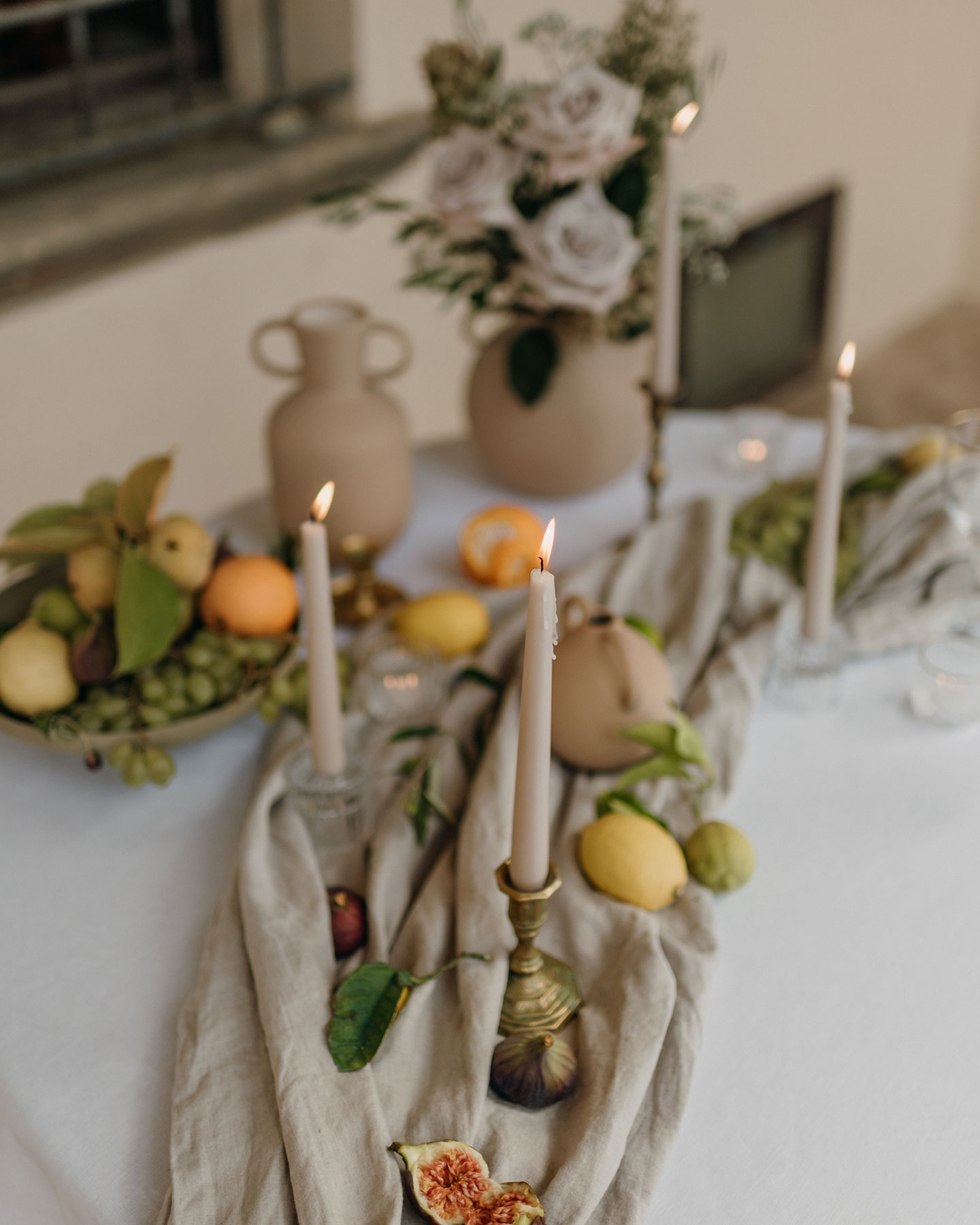 Der gedeckte Hochzeitstisch wurde mit einer weißen Tischdecke versehen. Darauf befindet sich ein Leinenläufer in Beige, auf diesem sind goldene Stabkerzenhalter, Zitrusfrüchte, Feigen und Dekoblätter platziert.